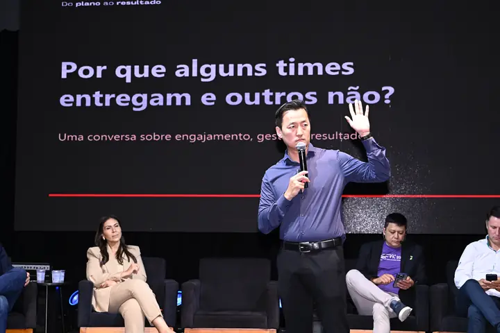 Palestrante gesticula no palco enquanto debatedores acompanham sentados