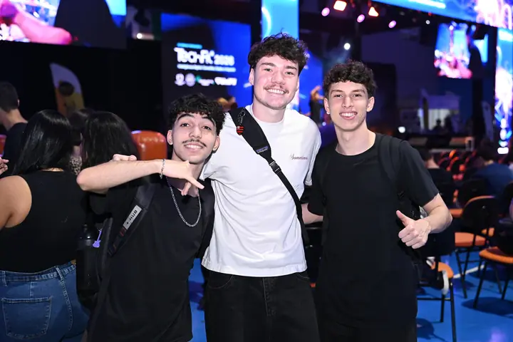 Três amigos sorriem para a câmera entre as cadeiras do evento