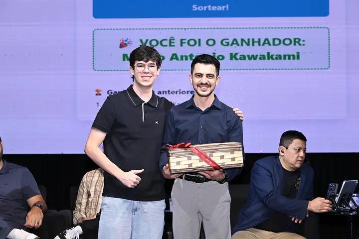 Ganhador do sorteio segura a cesta de prêmios diante do telão